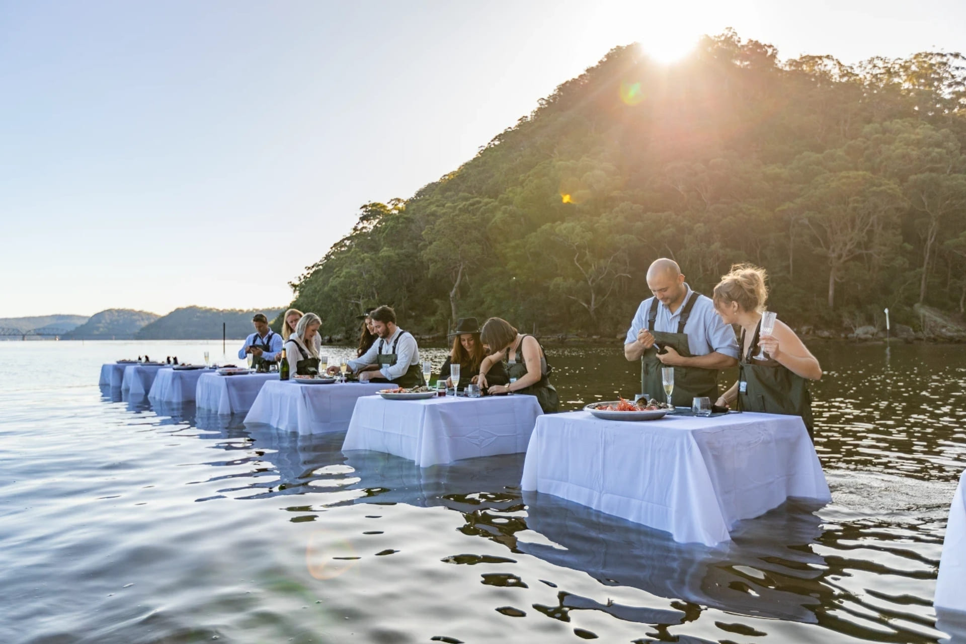 Guests enjoy a meal at tables set in the water beside Hawkesbury river shoreline at sunset.
